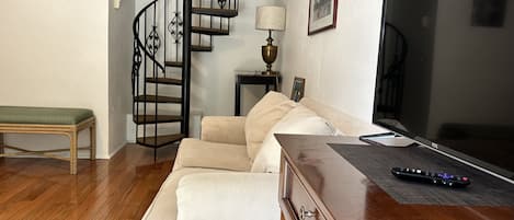 Una sala de estar con piso de madera, un sofá color beige, una mesa de centro de madera, un televisor, un control remoto, una lámpara y una escalera.