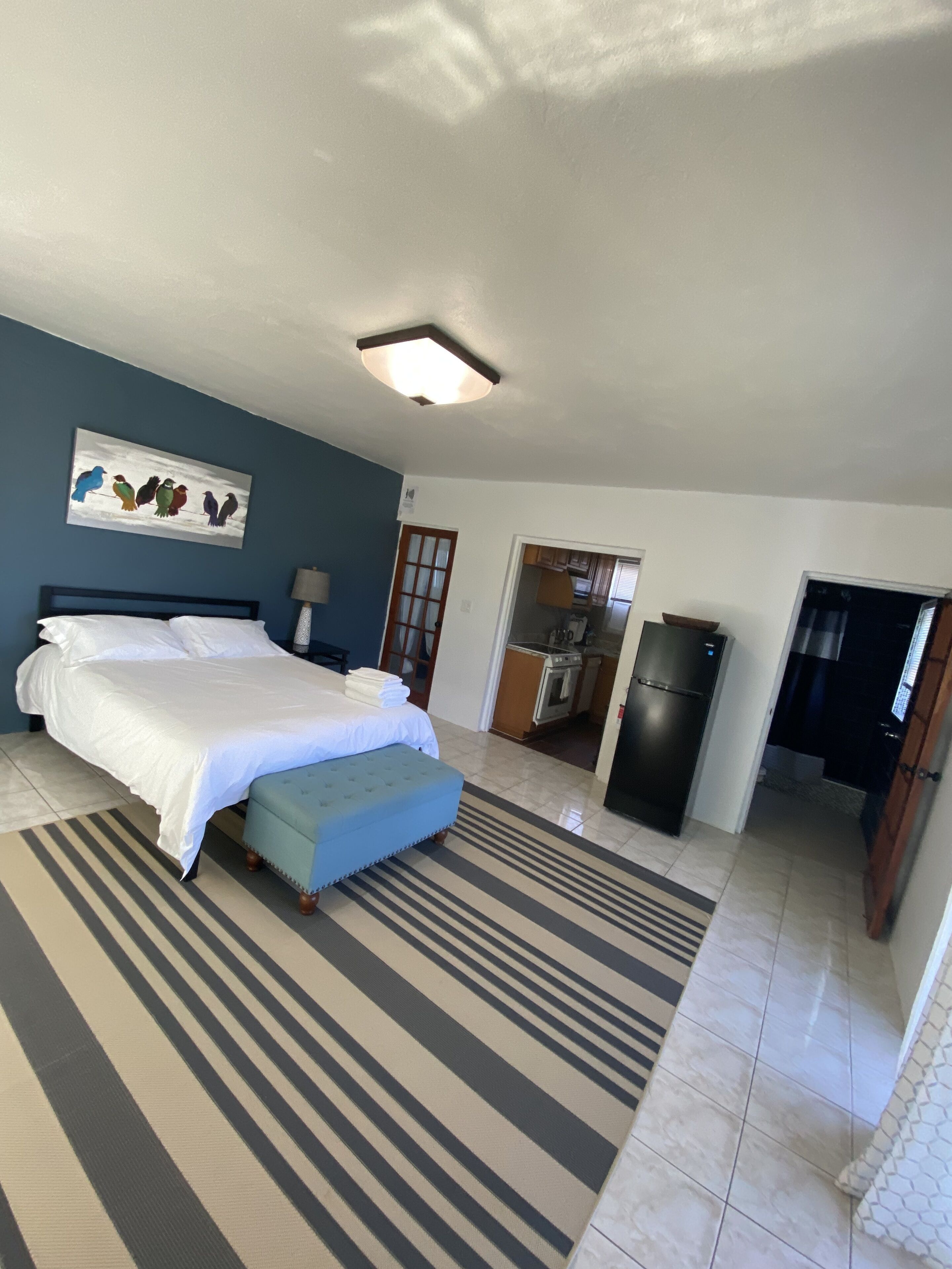 A bedroom with a bed, a bedside table, a lamp, a painting, and a view of a kitchen and dining area.