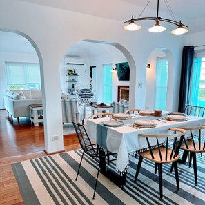 A dining area with a long table set for six, surrounded by chairs, in a room with white walls and wooden flooring.