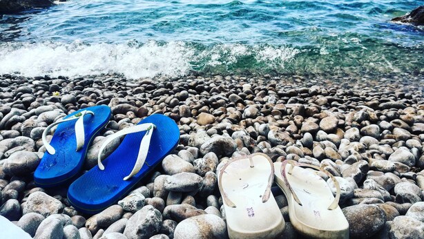 Une plage de galets où l’on aperçoit une paire de tongs bleues et une paire de tongs blanches.