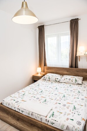 Una cama bien hecha con una colcha estampada, un cabecero de madera, una lámpara de noche y una ventana con cortinas.