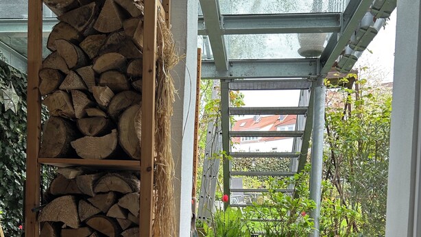 Un espace extérieur abrité comprenant une étagère en bois, une chaise en osier avec un coussin rose et une plante en pot.