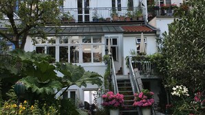 Un immeuble résidentiel à plusieurs étages, doté d’un jardin bien entretenu, d’un escalier apparent et d’un balcon agrémenté de plantes en pot.