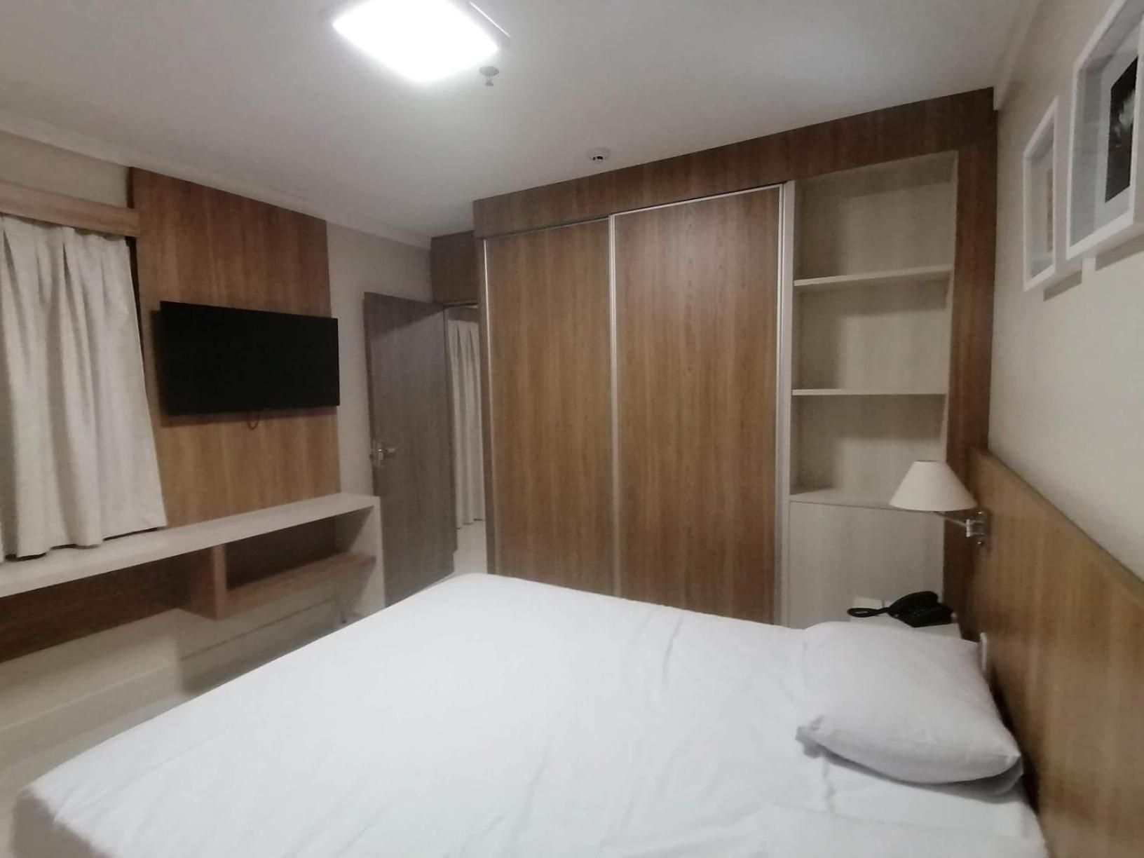 Un dormitorio con cama, televisor montado en la pared, armario y mesita de noche con lámpara.