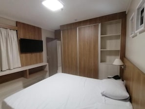 Un dormitorio con cama, televisor montado en la pared, armario y mesita de noche con lámpara.