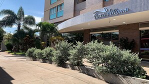 A hotel with a modern facade, a sign reading 'Piazza Roma,' and a landscaped area with various shrubs and trees.