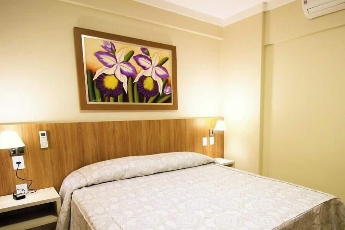 A hotel room with a bed, a nightstand, a lamp, a painting of flowers, and a wall-mounted air conditioner.
