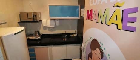 A kitchen with a microwave, stove, and a refrigerator. There is a wall-mounted television and a poster with the text 'COPA DA MAMÃE'.