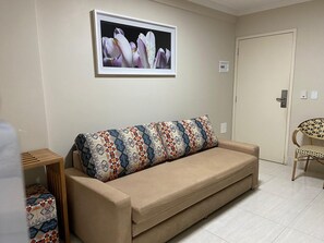 A beige sofa with patterned cushions, a framed picture on the wall, a small wooden table, and a wicker chair. - Piazza diRoma (Caldas Novas)
