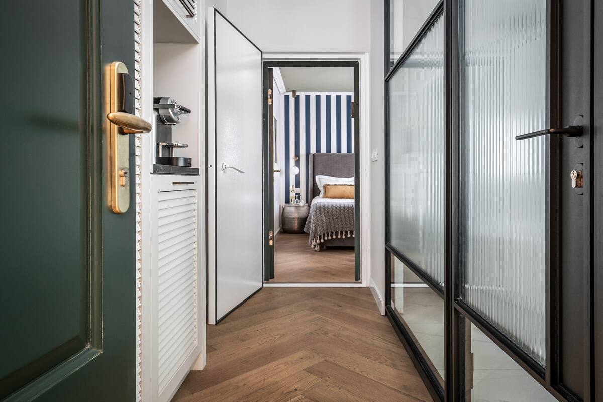 A hallway with a wooden floor leading to a bedroom with striped wallpaper and a bed with a gray headboard.