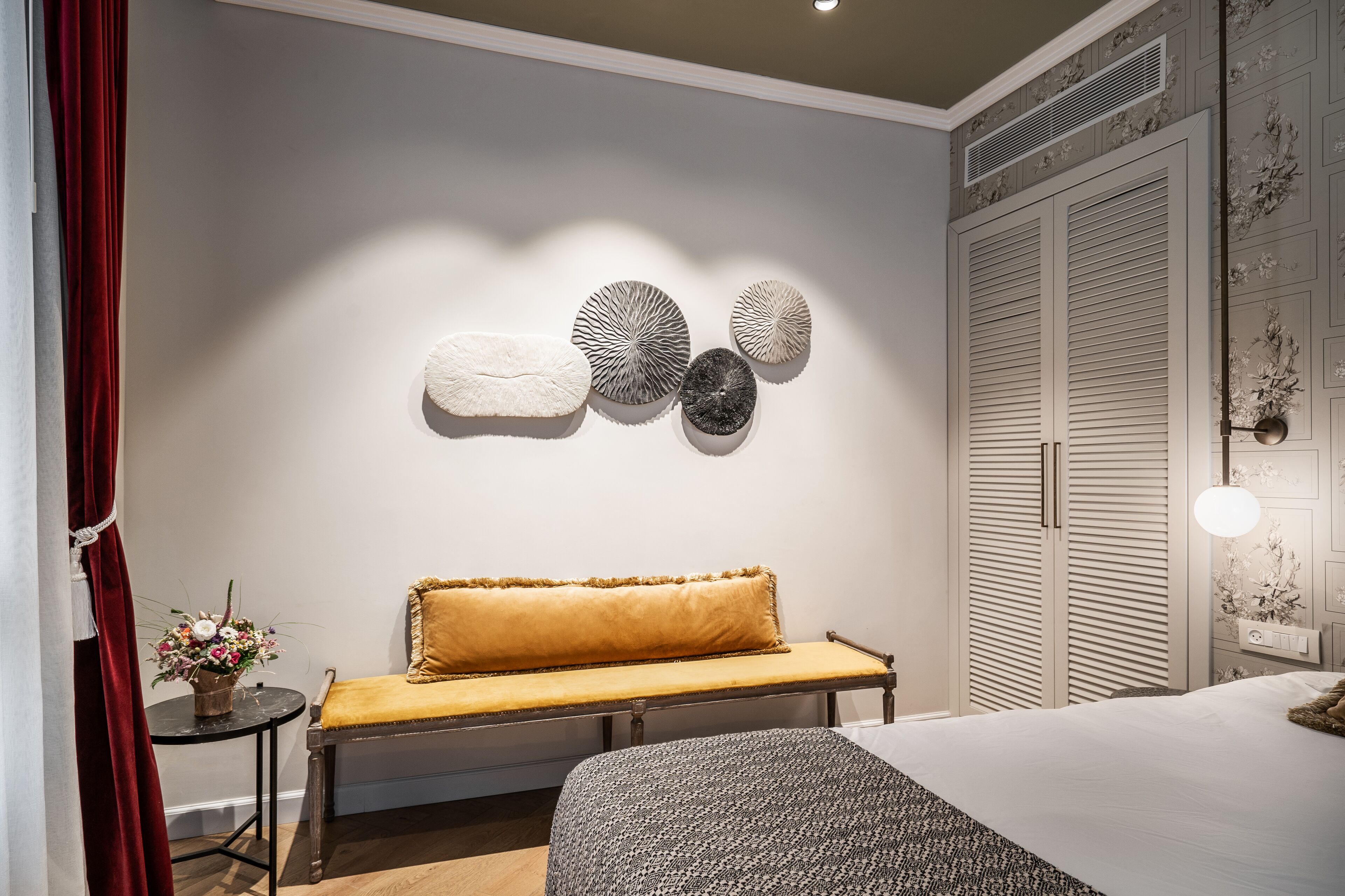 Une chambre d’hôtel avec un lit, un banc, une petite table sur laquelle est posé un vase de fleurs, et un mur décoré d’assiettes.