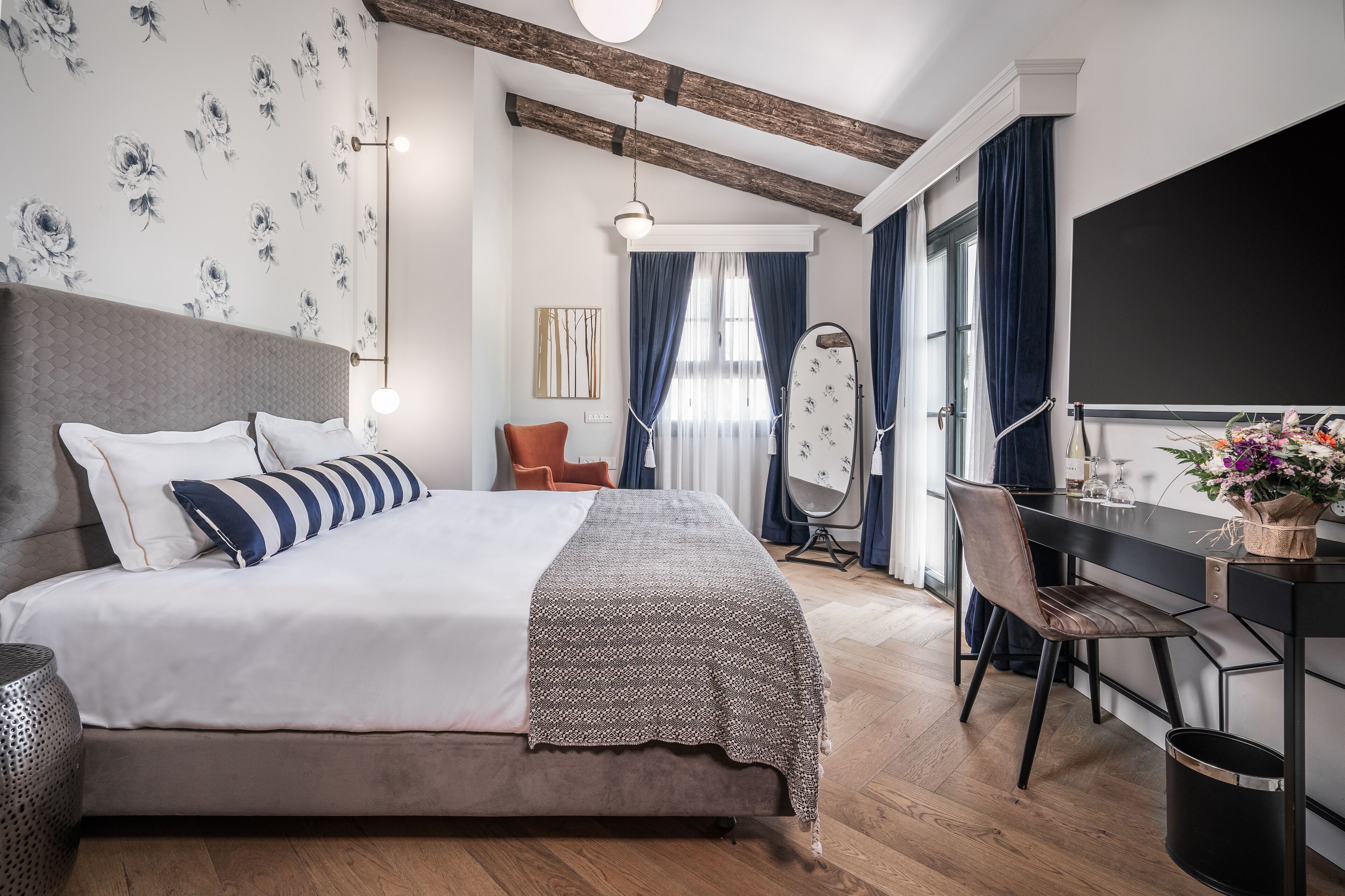 Habitación de hotel moderna con una cama grande, un escritorio y una silla.  Hay un televisor, una ventana con cortinas y una pared con estampado floral.
