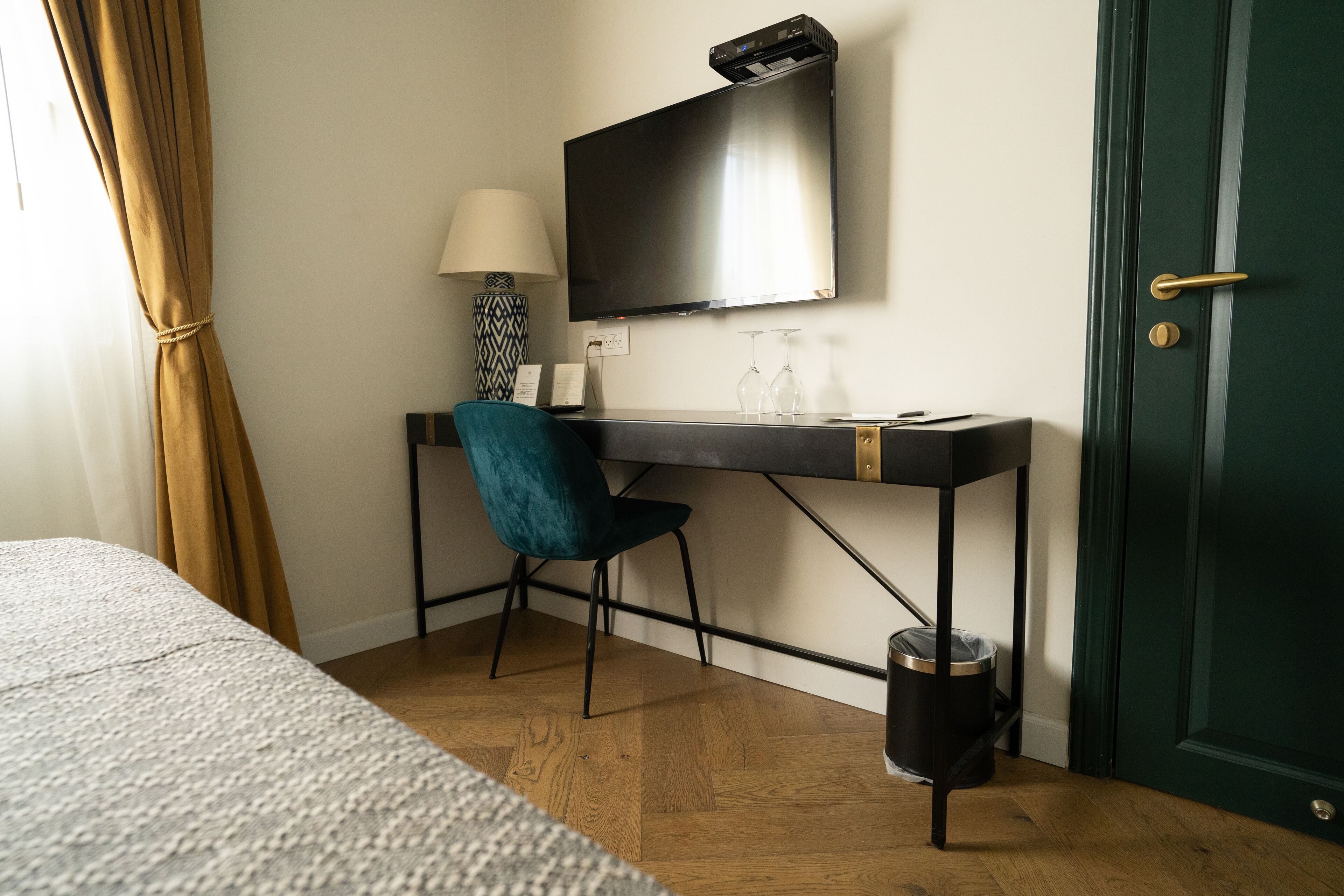 Una habitación de hotel con cama, escritorio con televisor, silla, lámpara y cesto de basura.