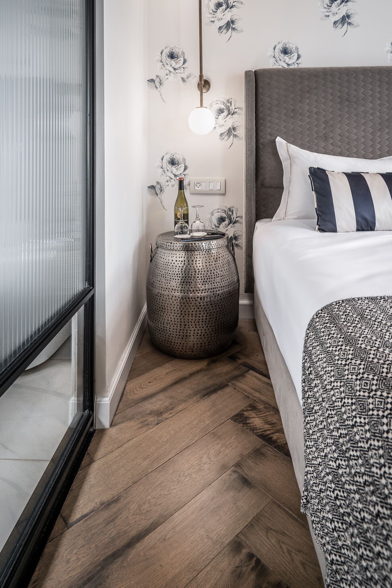 A bedroom with a bed, a round side table, a wall clock, and a floral wallpaper pattern.