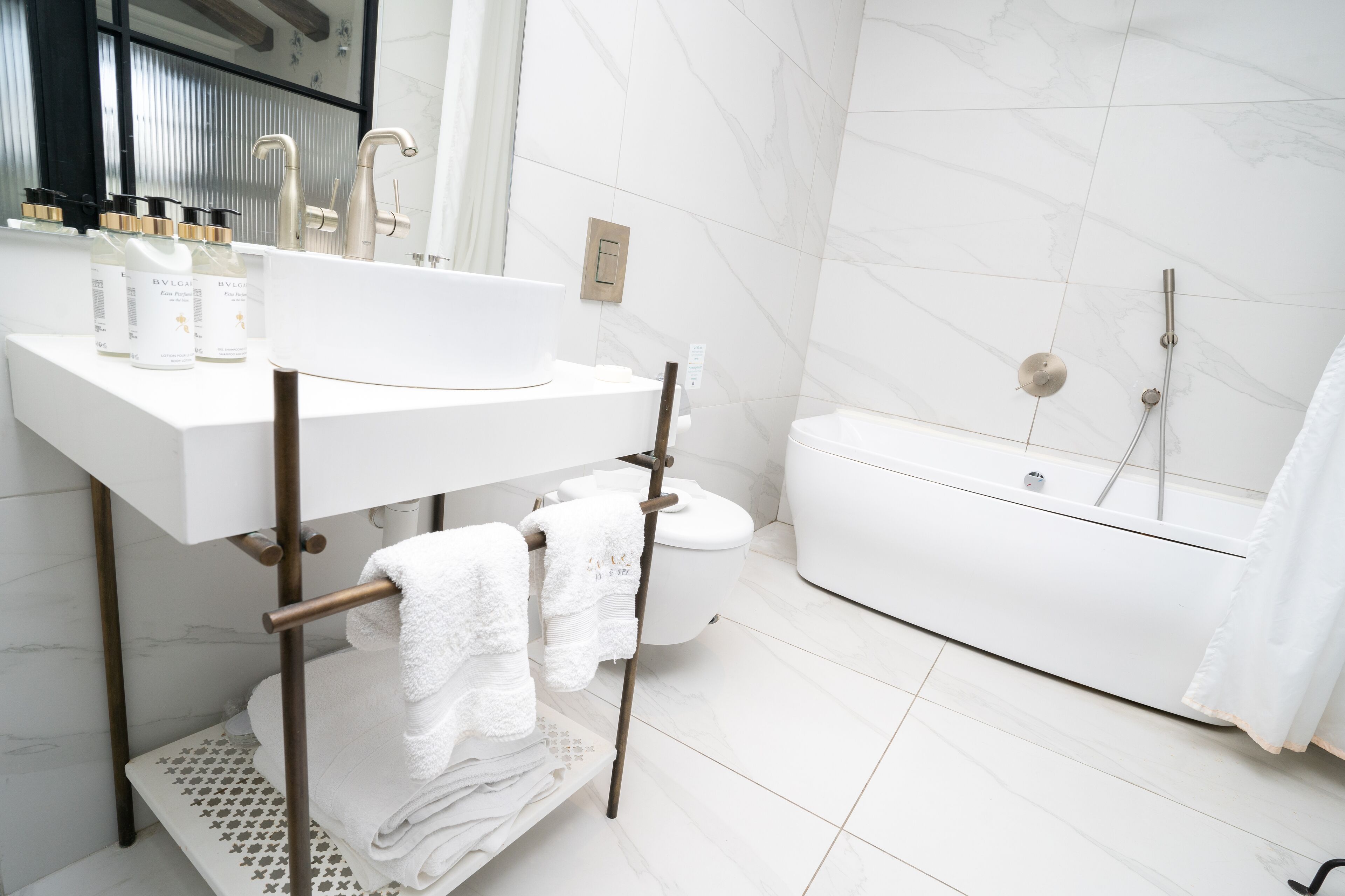 Un baño moderno con un lavamanos, tocador y bañera blancos. Hay toallas colgadas en un perchero y un inodoro visible al fondo.