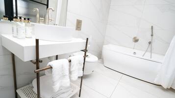 Un baño moderno con un lavamanos, tocador y bañera blancos. Hay toallas colgadas en un perchero y un inodoro visible al fondo.