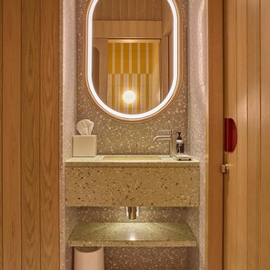 Un bagno con una porta in legno, uno specchio con luce, un lavandino e un dispenser di sapone, e un porta asciugamani.