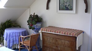 A room with a sloped ceiling, a wooden dresser, a small table with a purple tablecloth, a wicker chair, a blue and white vase with a plant, and a framed painting on the wall. - Beautiful bright room (Le Mesnil-Esnard)