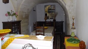 Une pièce avec un lit, une table recouverte d’une nappe jaune, un vase rempli de fleurs et un miroir accroché au mur.