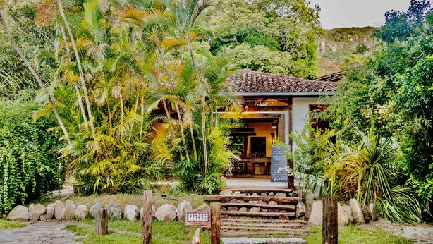 Un pequeño edificio rústico con tejado de baldosas, rodeado de abundante vegetación y un sendero de piedra que conduce a él.