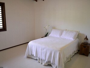 Quarto simples e limpo com uma cama, mesa de cabeceira, uma pequena mesa e uma janela.