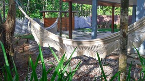 A hammock in a garden area with a wooden deck and a fence in the background. - Casa Palmeira Condomínio em Trancoso (Porto Seguro)