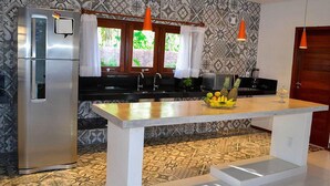A kitchen with a stainless steel refrigerator, a central island with a fruit bowl, a sink, and a window with curtains. - Casa Palmeira Condomínio em Trancoso (Porto Seguro)