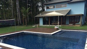 A rectangular swimming pool with a tiled edge, surrounded by a wooden deck with lounge chairs. - Casa Palmeira Condomínio em Trancoso (Porto Seguro)