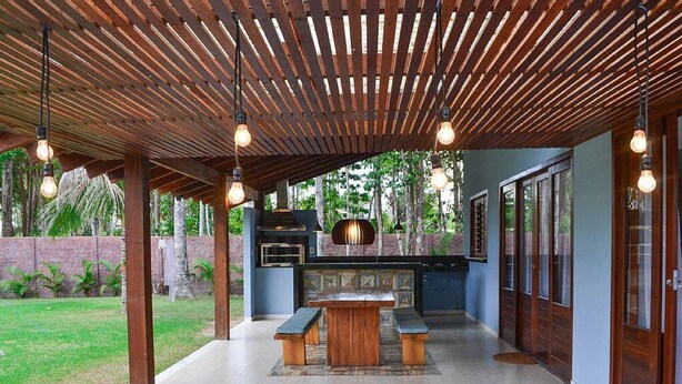 An outdoor kitchen area with a built-in oven, a wooden table, and hanging light fixtures. - Casa Palmeira Condomínio em Trancoso (Porto Seguro)