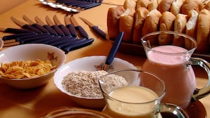 Una mesa con tazones de cereal, un jarro de batido rosa y una bandeja de panecillos.