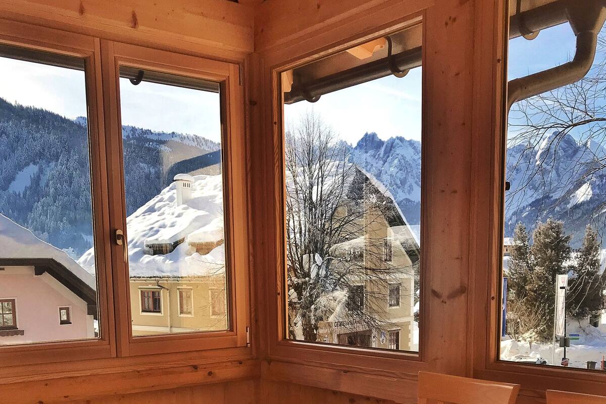 A wooden interior with a dining table and chairs, large windows with a snowy mountain view.