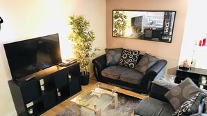 A living room with a flat-screen TV, a black sofa, a glass coffee table, and a potted plant. - Granary Mews (Crediton)