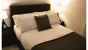 A neatly made bed with white linens and a dark throw blanket, flanked by two bedside lamps on matching nightstands. - Granary Mews (Crediton)