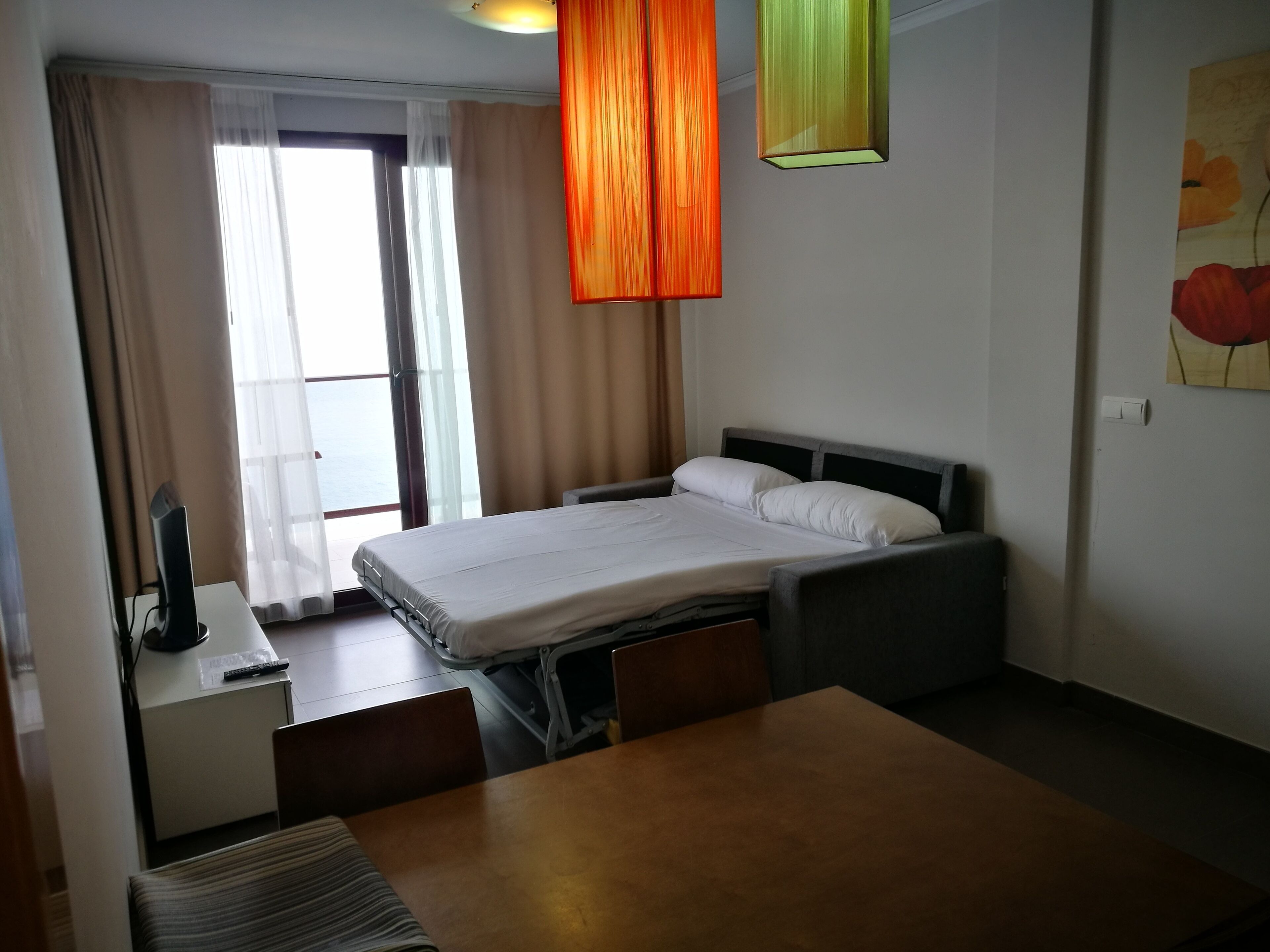 A hotel room with a bed, bedside table, and a desk with a chair. There is a window with curtains, a painting on the wall, and a hanging light fixture.