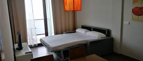 Habitación de hotel con cama, mesita de noche y escritorio con silla. Se observa una ventana con cortinas, un cuadro en la pared y una lámpara colgante.
