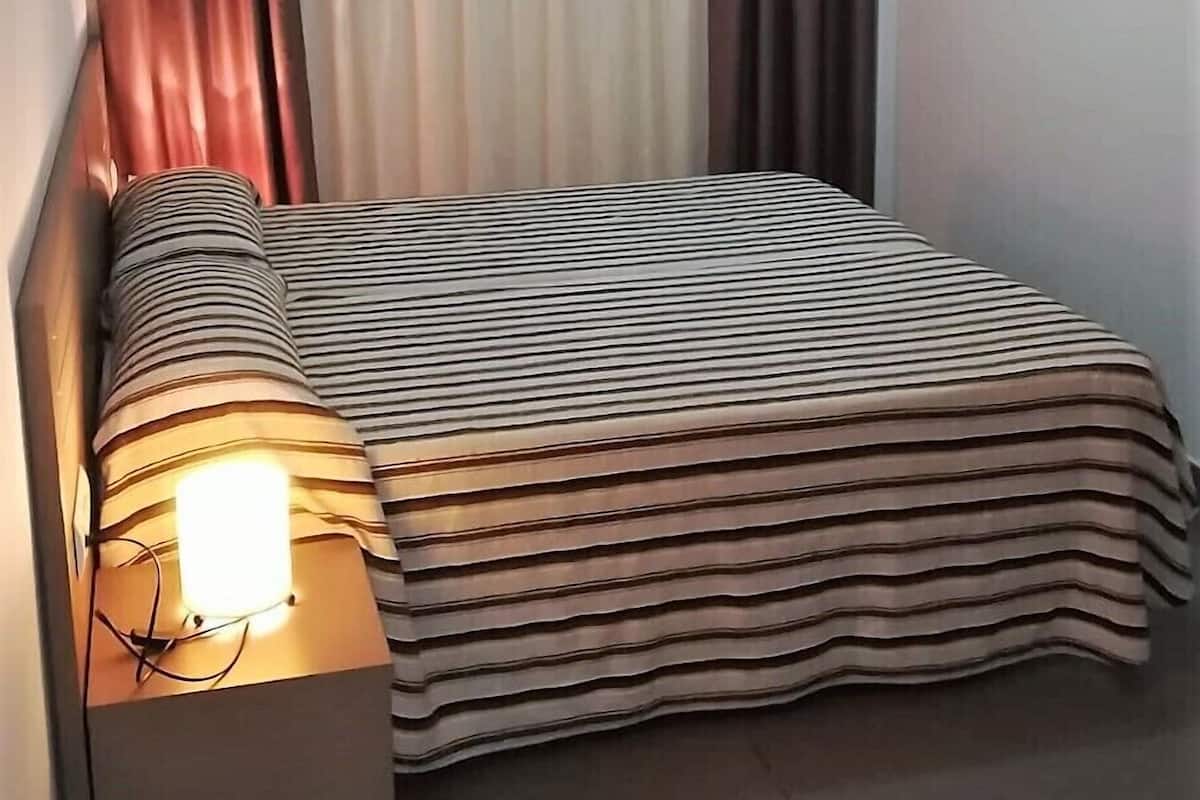 A small, neatly arranged bedroom with a bed, a bedside lamp, and a window with curtains.