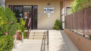 An entrance to a hotel named "APARTHOTEL EL FARO" with a sign indicating a 4-star rating. There are steps leading to the entrance, potted plants, and a metal fence on the right. - Aparthotel El Faro (Cullera)