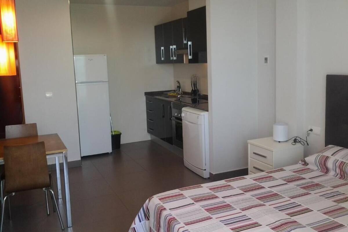 A compact hotel room with a bed, a small kitchenette, a dining area, and a bedside table with a lamp.