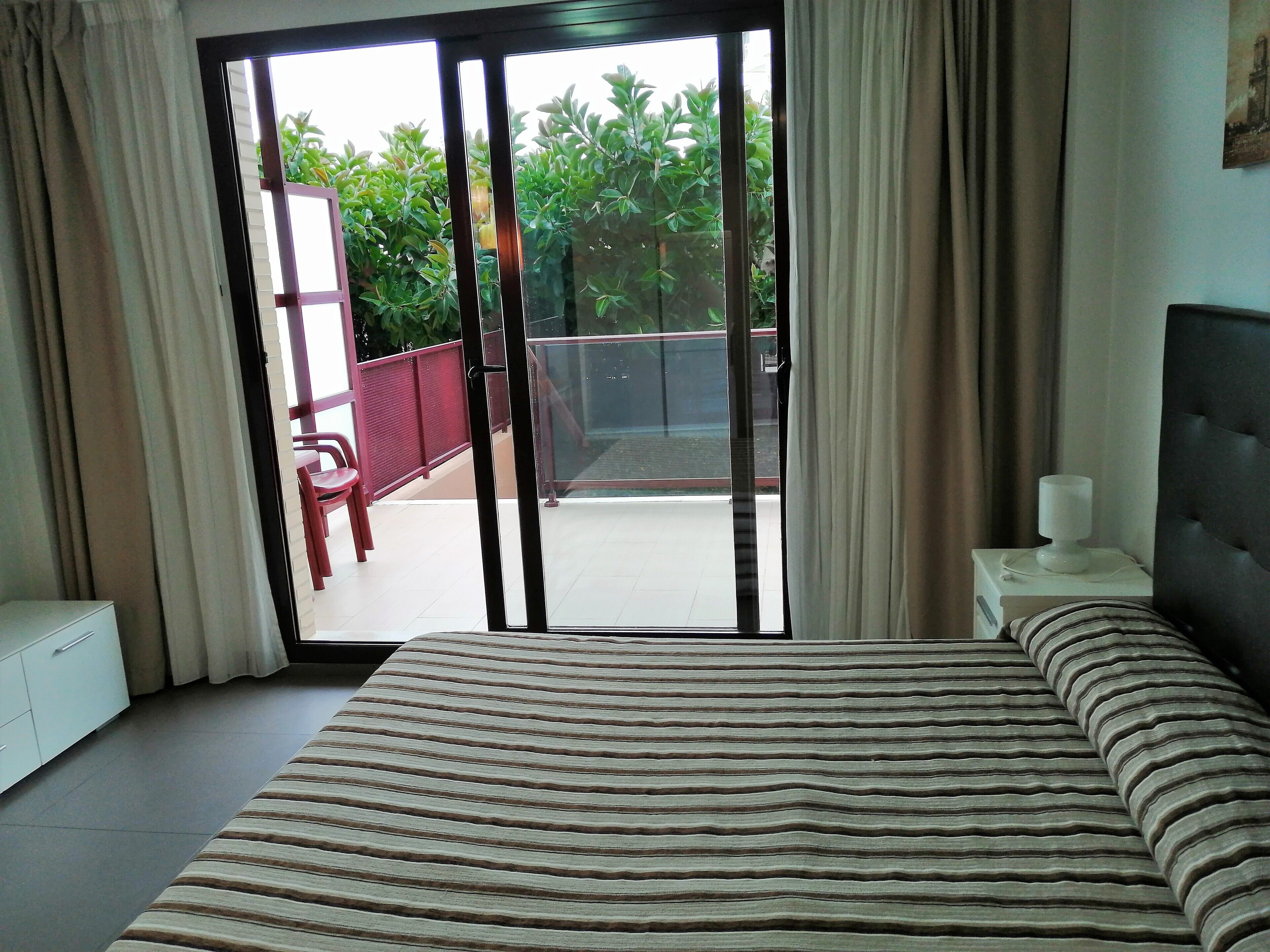 Quarto de hotel com cama, mesa de cabeceira, janela com cortinas e vista para uma varanda e vegetação lá fora.