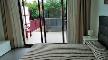 Quarto de hotel com cama, mesa de cabeceira, janela com cortinas e vista para uma varanda e vegetação lá fora.