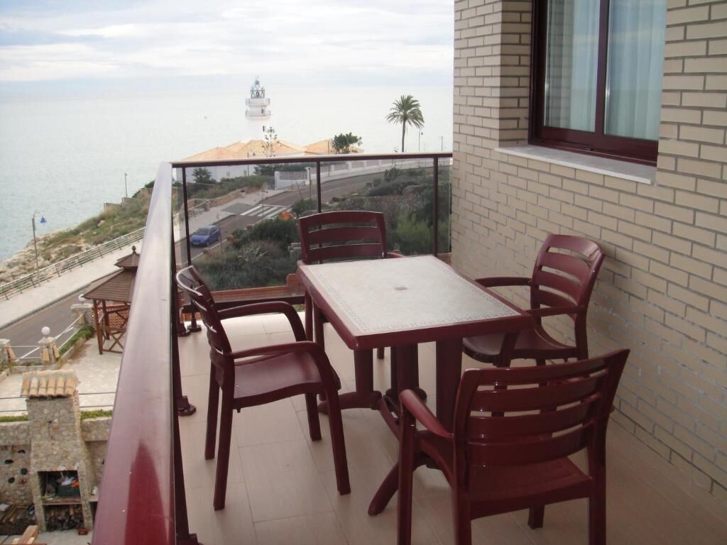 A balcony with a table and chairs, overlooking a coastal road and a lighthouse.
