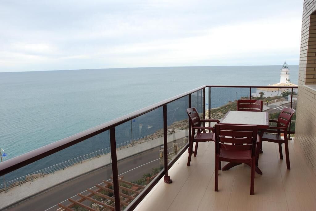 A balcony with a table and chairs overlooking the sea, a road, and a lighthouse.