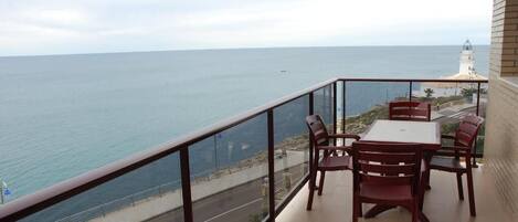 A balcony with a table and chairs overlooking the sea, a road, and a lighthouse.