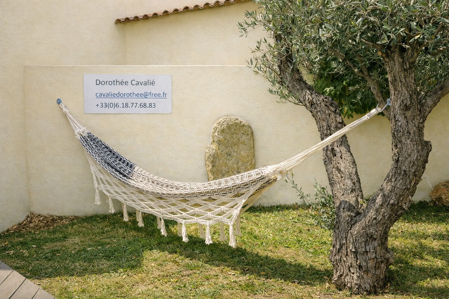 A hammock tied between two trees, a stone wall, and a sign with contact information.