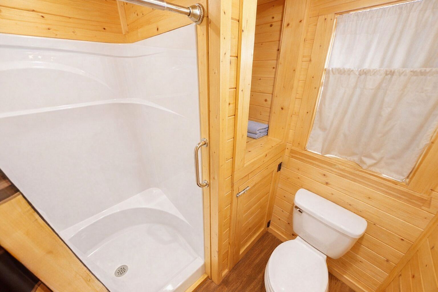 A wooden cabin bathroom with a shower, toilet, and a window with a curtain.