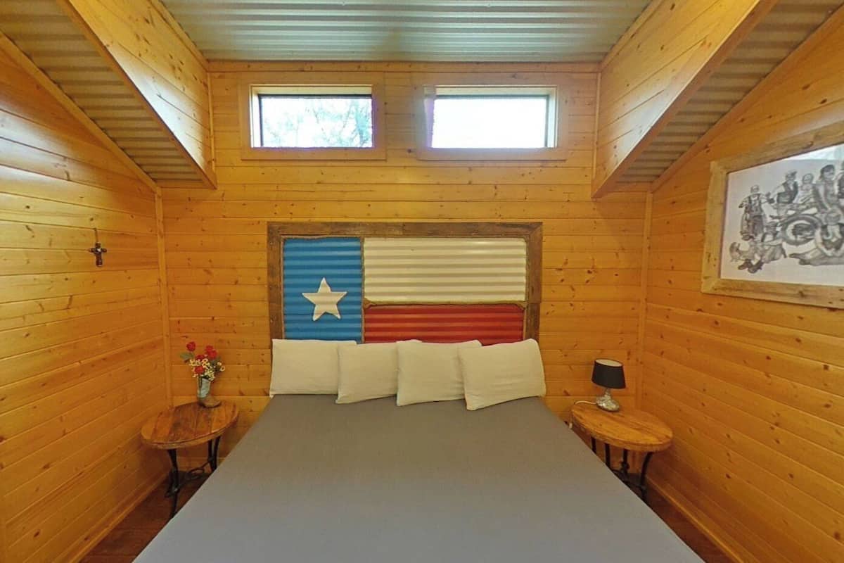A wooden cabin bedroom with a bed, two windows, a wall-mounted picture, and a small table with a lamp.