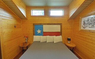 A wooden cabin bedroom with a bed, two windows, a wall-mounted picture, and a small table with a lamp.