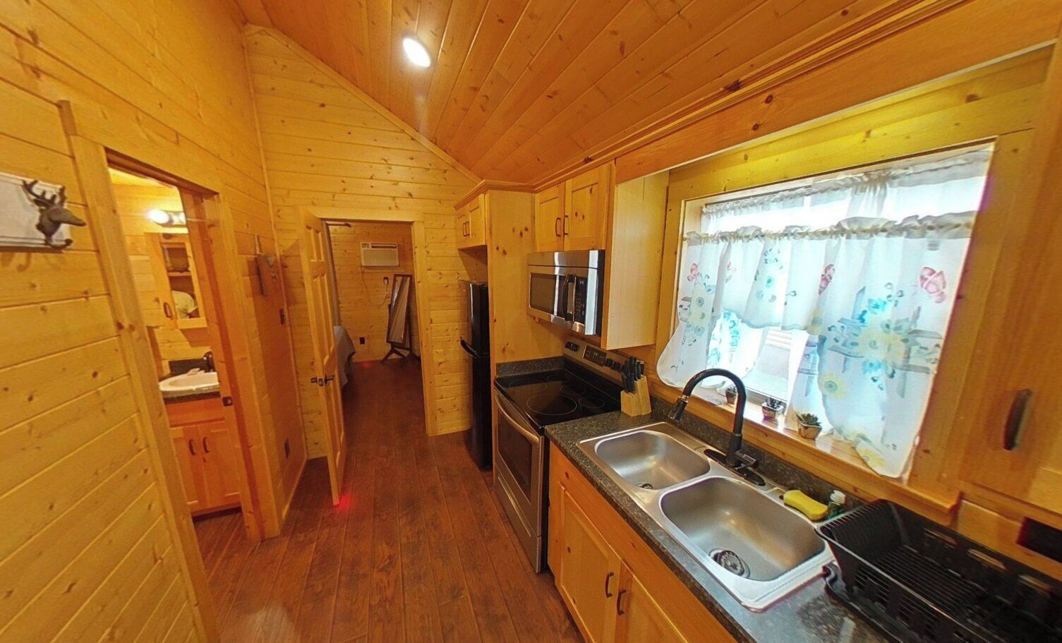 A wooden cabin interior with a kitchen, wooden walls, and a window with a curtain.
