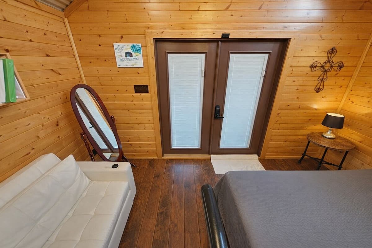 A wooden-paneled room with a bed, a chair, a small table with a lamp, and a door with a sign on the wall.