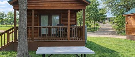 A wooden cabin with a porch, a picnic table, and a view of the surrounding greenery.